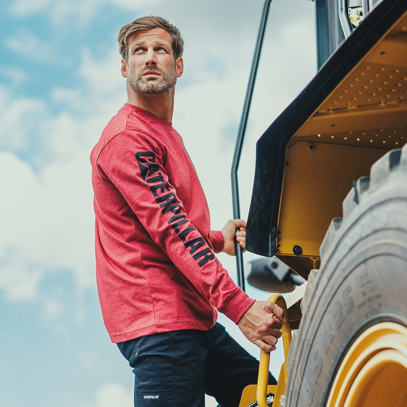 Man climbing up plant machinery, wearing a Caterpillar Trademark Banner Long Sleeve T-Shirt in brick